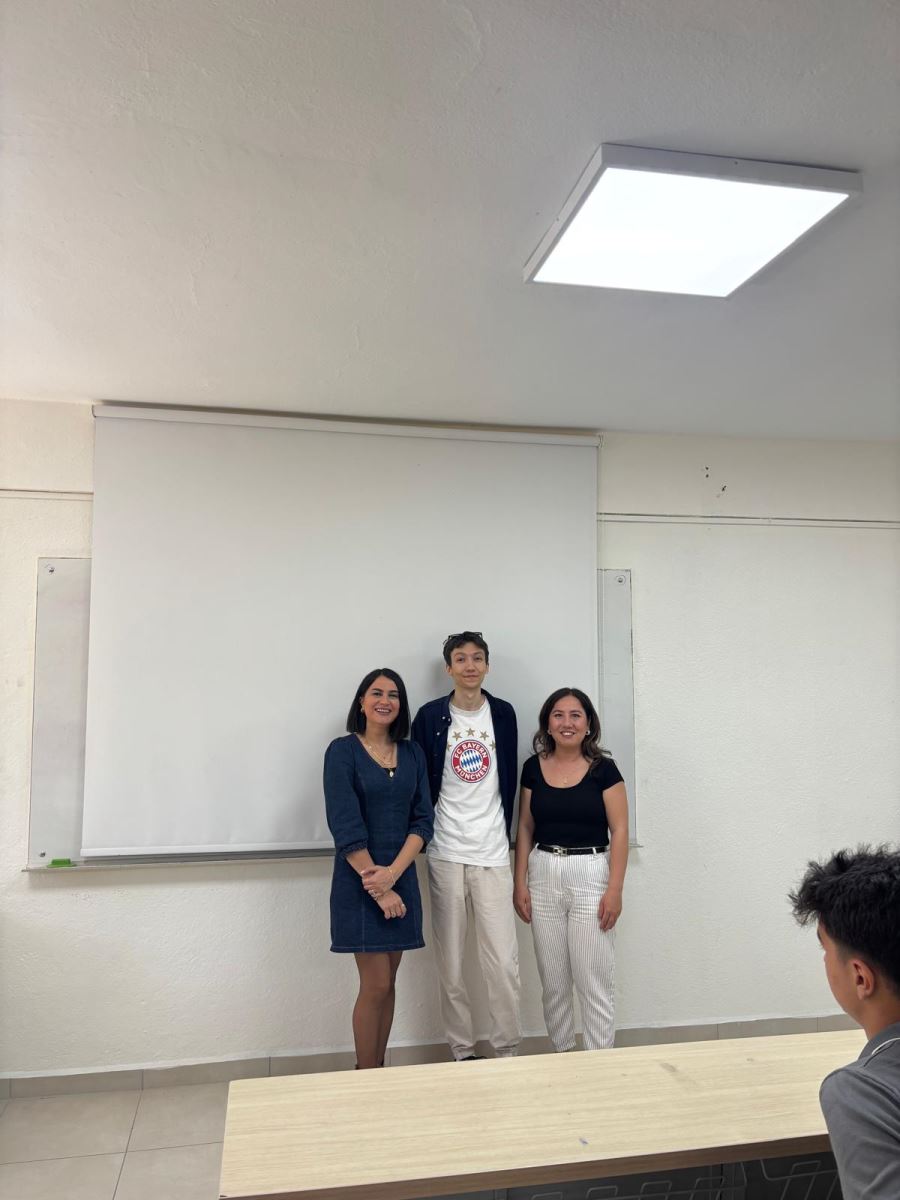 A photo taken in a classroom shows three people standing side by side in front of a white projection screen. There is a row of light-colored desks in the foreground. A square-shaped light fixture is mounted on the classroom ceiling. The walls and the classroom setting have a simple, uncluttered appearance.